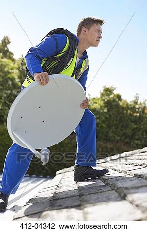Stock Image - Worker installing satellite dish on roof. Fotosearch