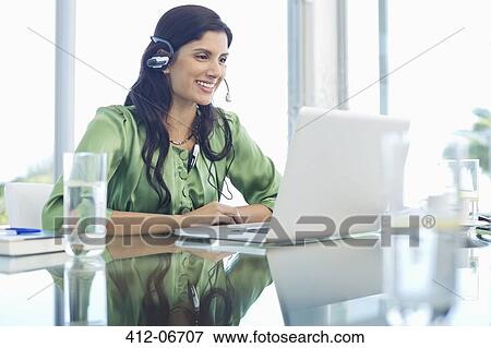 Stock Photo - Businesswoman listening to headphones at desk. Fotosearch