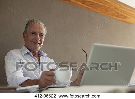 Stock Image - Older man having cup of coffee at desk. Fotosearch