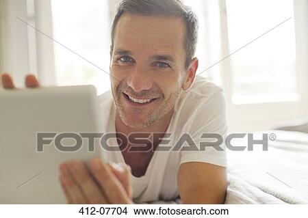 Picture - Portrait of smiling man using digital tablet in bed. Fotosearch