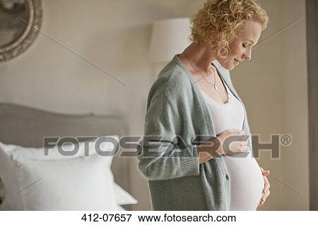 Stock Photo - Smiling pregnant woman touching stomach in bedroom. Fotosearch