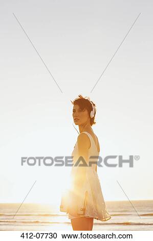 Stock Image - Woman in dress wearing headphones on sunny beach. Fotosearch