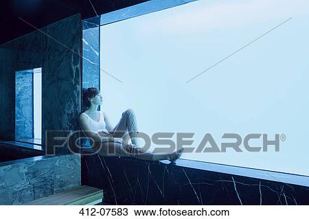 Stock Image - Woman sitting on window ledge and looking at underwater view of swimming pool. Fotosearch