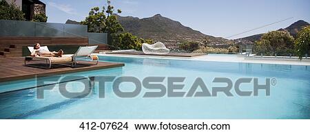 Picture - Woman sunbathing on lounge chair next to luxury swimming pool with mountain view. Fotosearch