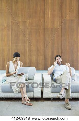 Stock Photo - Couple using laptop and cell phone in armchairs. Fotosearch