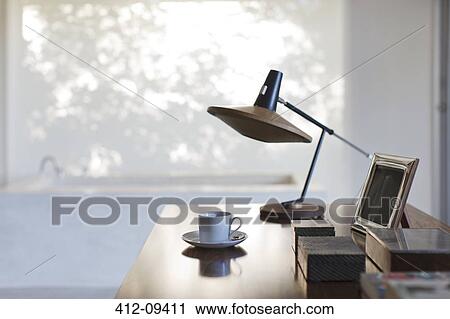 Stock Image - Cup of coffee and lamp on desk in office. Fotosearch