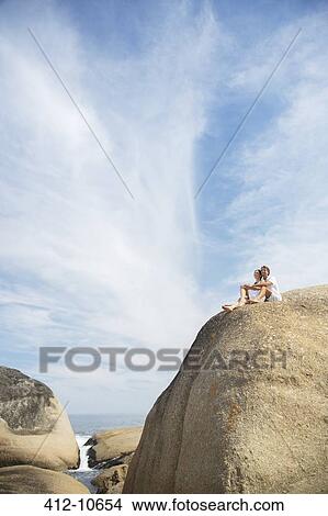 Picture - Couple sitting on rock formation on beach. Fotosearch