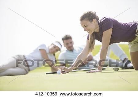 Stock Photo - Friends watching woman flick golf ball toward hole. Fotosearch