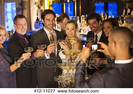 Stock Photo - Man photographing bride and groom with friends at champagne pyramid. Fotosearch
