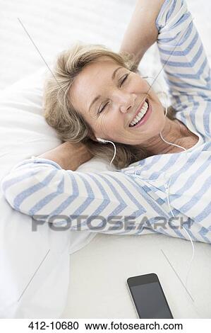 Stock Image - Older woman listening to earphones on bed. Fotosearch