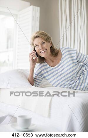 Stock Image - Older woman talking on cell phone on bed. Fotosearch