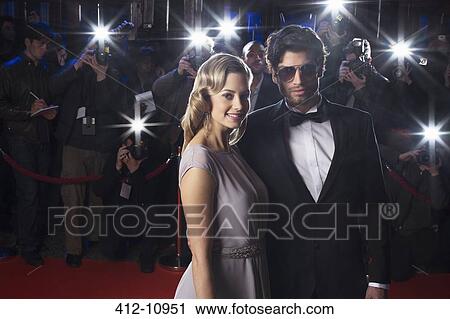 Stock Image - Serious celebrity couple on red carpet with paparazzi in background. Fotosearch