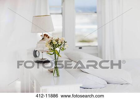 Stock Photograph - Vase of flowers and lamp in white bedroom. Fotosearch