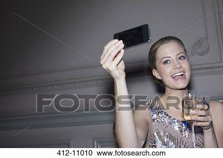 Stock Image - Well dressed woman with champagne taking self-portrait with camera phone. Fotosearch