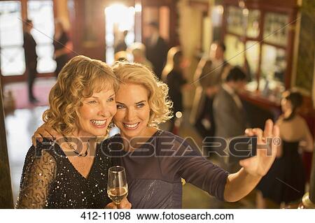 Stock Photo - Well dressed women with champagne taking self-portrait with camera phone in theater lobby. Fotosearch