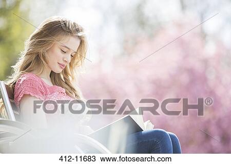 Stock Image - Woman reading book on park bench. Fotosearch