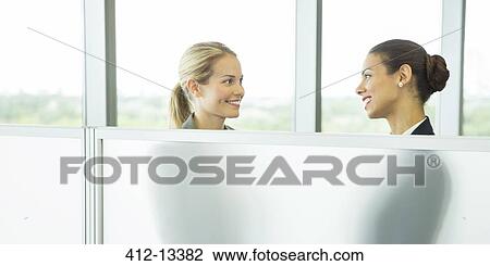 Stock Image - Businesswomen talking behind half wall in office. Fotosearch