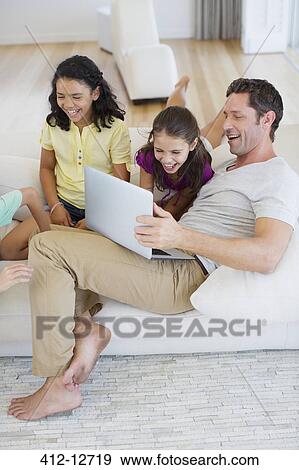 Stock Photo - Father and daughters using laptop on sofa in living room. Fotosearch