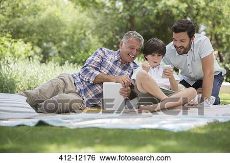 Stock Photograph - Multi-generation men with cell phone on blanket in grass. Fotosearch