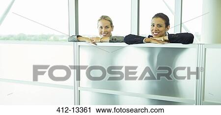 Stock Image - Smiling businesswomen leaning against half wall in office. Fotosearch