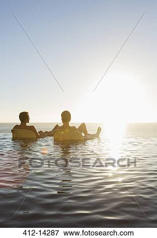 Stock Photo - Couple in lawn chairs in infinity pool overlooking ocean. Fotosearch