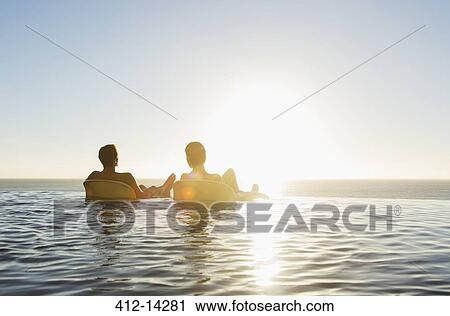 Stock Image - Couple in lounge chairs in infinity pool overlooking ocean. Fotosearch