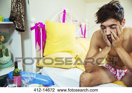 Stock Image - Hungover man rubbing face on bed the morning after a party. Fotosearch