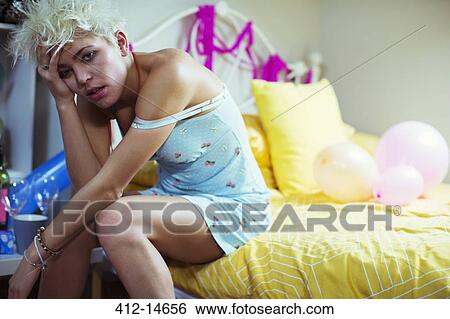 Stock Photograph - Hungover woman sitting on bed the morning after a party. Fotosearch