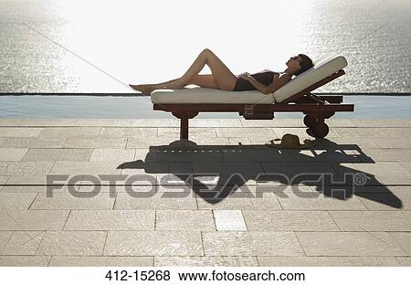 Stock Photo - Woman sunbathing in lounge chair at poolside overlooking ocean. Fotosearch