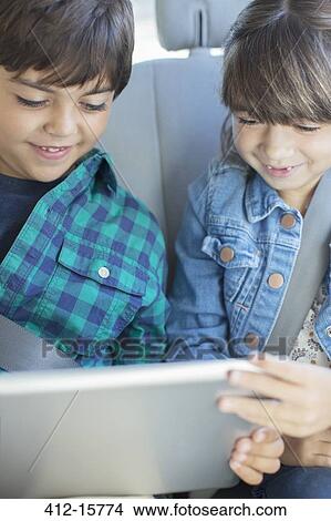 Picture - Brother and sister sharing digital tablet in back seat of car. Fotosearch