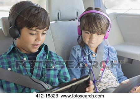 Stock Photo - Brother and sister with headphones using digital tablets in back seat of car. Fotosearch