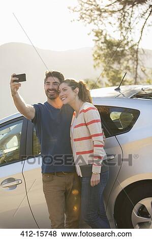 Stock Photo - Couple taking self-portrait with camera phone outside car. Fotosearch