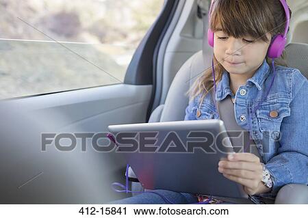 Stock Image - Girl with headphones using digital tablet in back seat of car. Fotosearch