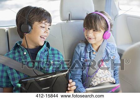 Picture - Happy brother and sister with headphones using digital tablets in back seat of car. Fotosearch