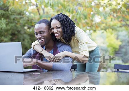 Stock Image - Happy couple using laptop at patio table. Fotosearch