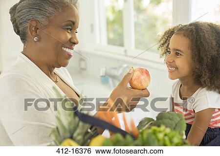 Happy grandmother giving apple to granddaughter in kitchen View Large Photo Image Stock Photograph - Happy grandmother giving apple to granddaughter in kitchen. Fotosearch