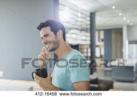 Stock Photo - Happy man talking on cell phone and drinking coffee at home. Fotosearch