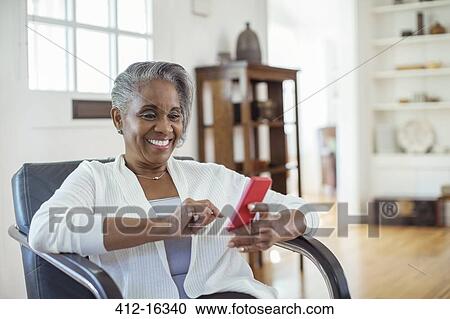 Stock Image - Happy senior woman text messaging with cell phone in living room. Fotosearch
