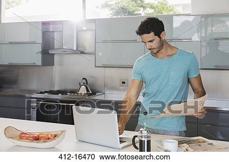 Stock Image - Man with newspaper using laptop in kitchen. Fotosearch