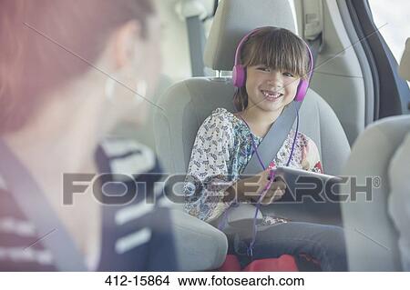 Picture - Mother turning and smiling at daughter with headphones and digital tablet in back seat of car. Fotosearch