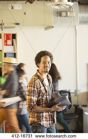 Stock Image - Portrait of confident casual businessman in office with co-workers passing by in background. Fotosearch