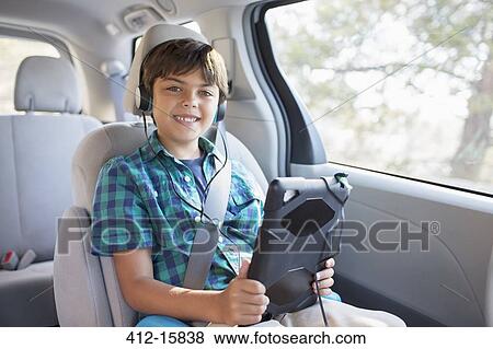 Stock Photo - Portrait of happy boy with headphones using digital tablet in back seat of car. Fotosearch