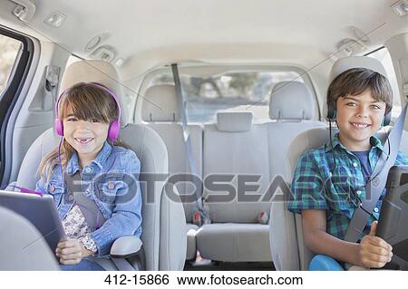 Stock Photograph - Portrait of happy brother and sister with headphones using digital tablets in back seat of car. Fotosearch