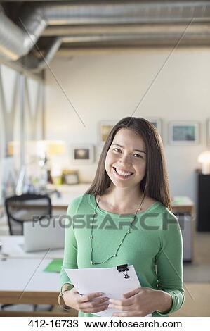 Stock Image - Portrait of smiling businesswoman holding paperwork in office. Fotosearch