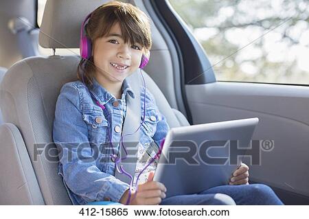 Stock Photography - Portrait of smiling girl with headphones using digital tablet in back seat of car. Fotosearch
