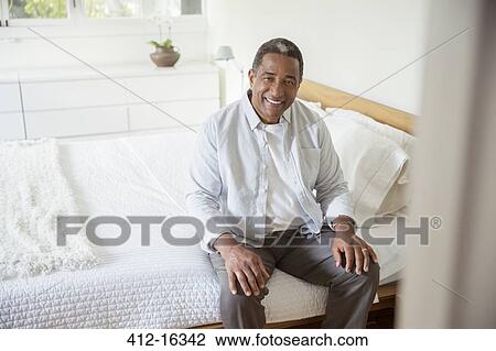 Stock Image - Portrait of smiling senior man sitting on bed. Fotosearch