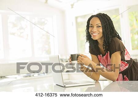 Stock Photography - Portrait of smiling woman drinking coffee at laptop in kitchen. Fotosearch