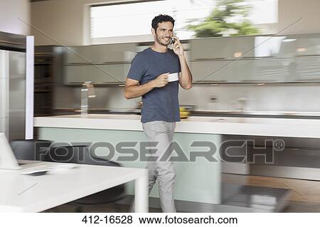 Stock Photo - Smiling man drinking coffee and talking on cell phone in kitchen. Fotosearch