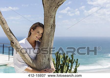 Stock Photo - Smiling woman in bathrobe hugging tree in front of ocean. Fotosearch