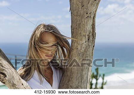 Stock Image - Wind blowing woman's hair in face behind tree trunk. Fotosearch